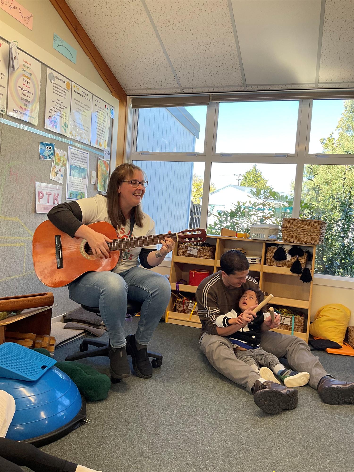 Joanna playing guitar in a music session while a child, supported by a caregiver, holds a rhythm stick
